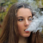 0_Vape-teenager-Young-pretty-white-girl-in-white-cardigan-smoking-an-electronic-cigarette-opposite-d.avif