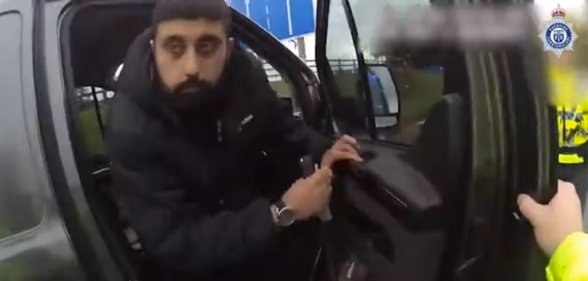 An individual, dressed in a dark jacket and sporting a beard, is seated inside a vehicle, appearing to handle the door handle. The scene suggests the person may be either entering or exiting the vehicle. The image is slightly blurred, providing a sense of movement or action.