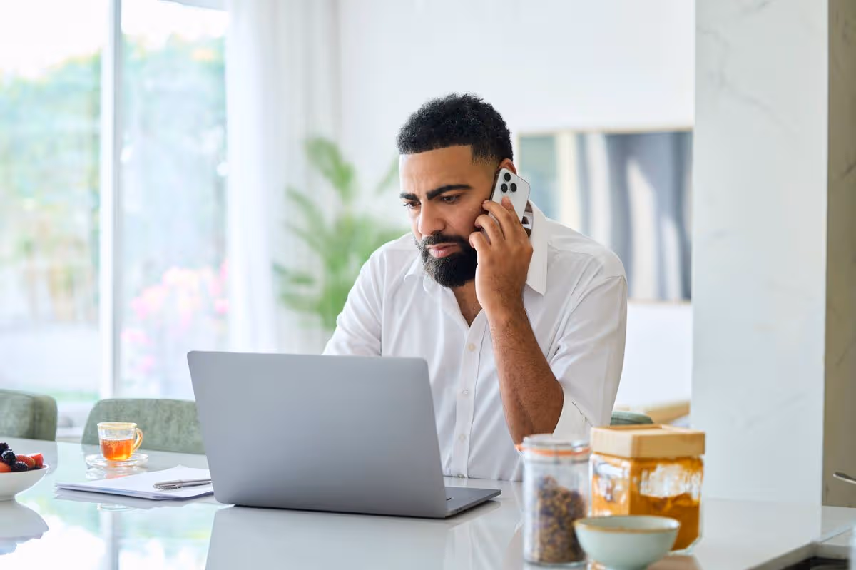 Middle eastern man talking on smart phone while looking at laptop. Emirati male is multi-tasking. He seems to be worried.