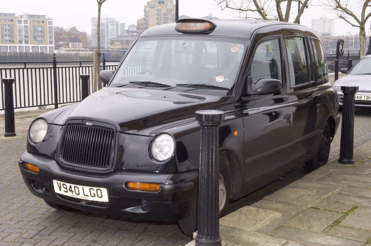 A black vehicle, possibly a taxi, is parked on the side of the road. The vehicle features a license plate with the word "LONDON" on it. The road is lined with bollards, and a portion of another vehicle is visible in the background.