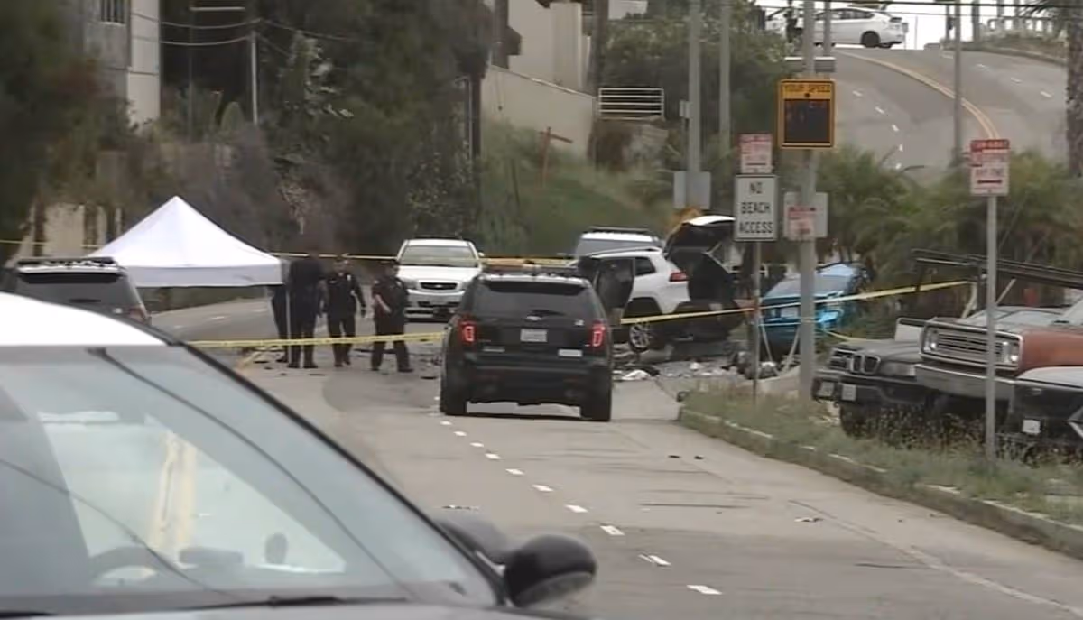 Urban street scene depicting a road closure with emergency vehicles and personnel present. A white tent is visible, and caution tape marks off a section of the road. Several cars are parked nearby, and a building can be seen in the background.