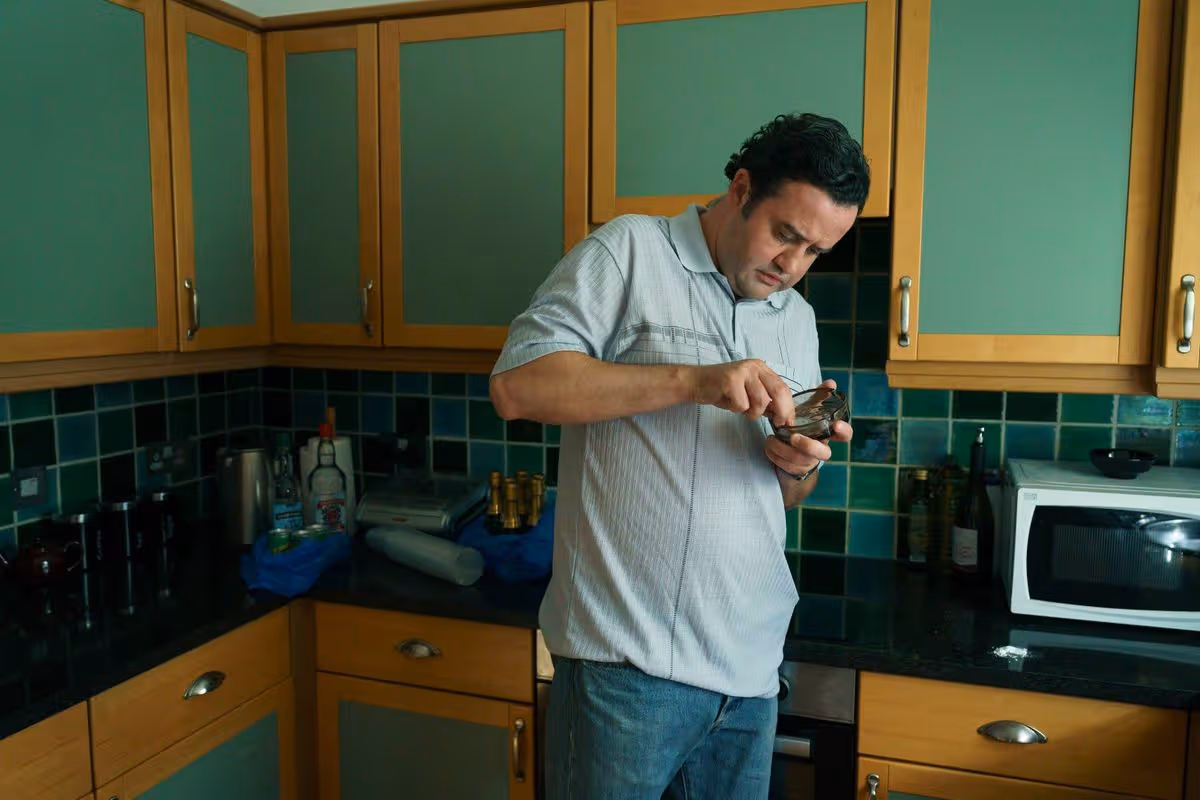 An individual standing in a kitchen, engaged in an activity that appears to involve a small electronic device, surrounded by kitchen cabinets and countertop.