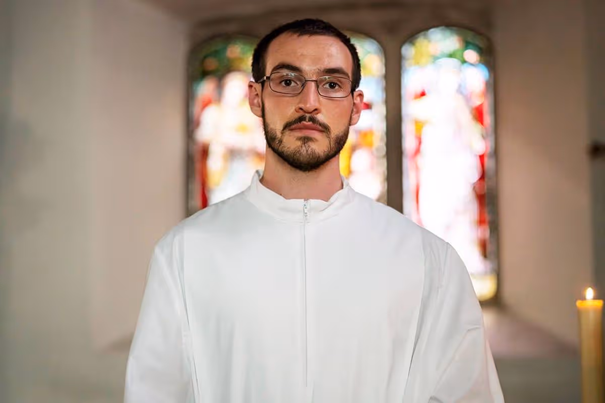 A man in a white shirt stands in front of a stained glass window