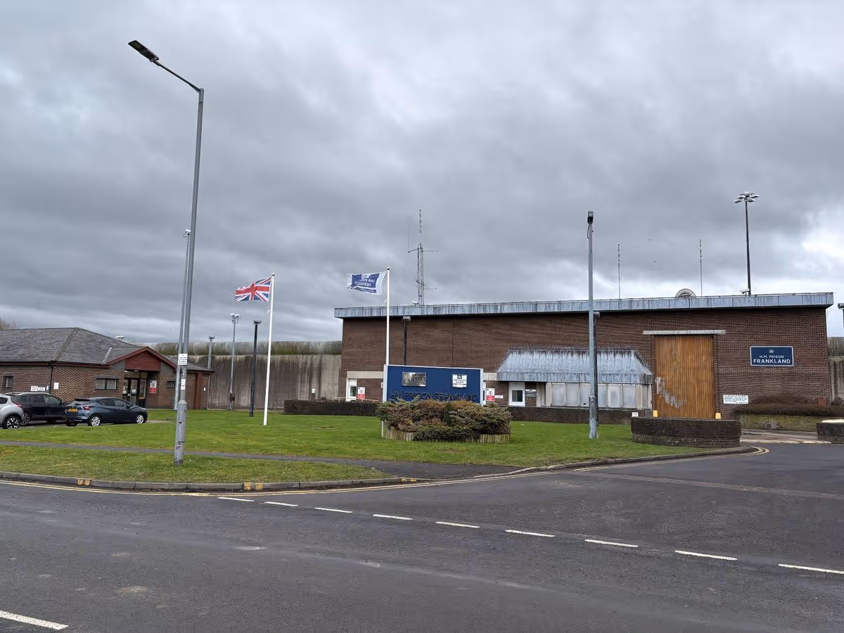 An image of an industrial building with several flags hoisted on poles in front. The building is surrounded by a grassy area with a road leading to it. The sky is cloudy and there is a vehicle parked nearby.