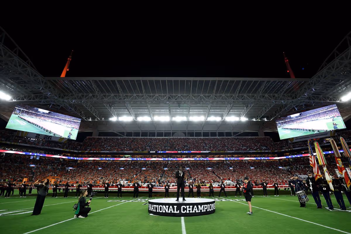 An individual is standing on a podium in the center of a sports stadium, with a large crowd gathered to watch. The field is marked with a prominent sign reading "National Champion."