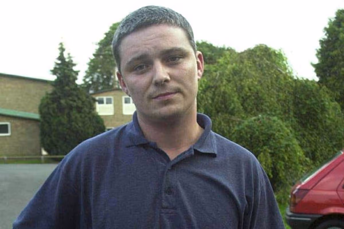 An individual dressed in a blue polo shirt is standing in an outdoor setting, with a red vehicle visible to his right and a backdrop of trees and a building.