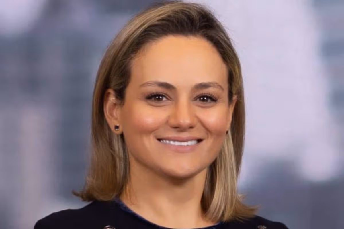 A close-up portrait of an individual with short blonde hair, wearing a black top, and smiling against a blurred background, likely in a professional setting.