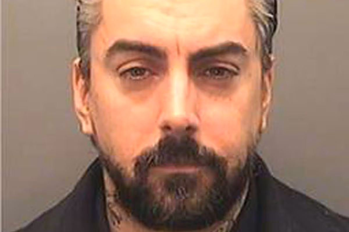 An individual, dressed in a dark shirt, is featured in a close-up photograph. This person exhibits a neatly groomed beard and mustache, along with a prominent furrow in the brow, standing against a plain background.