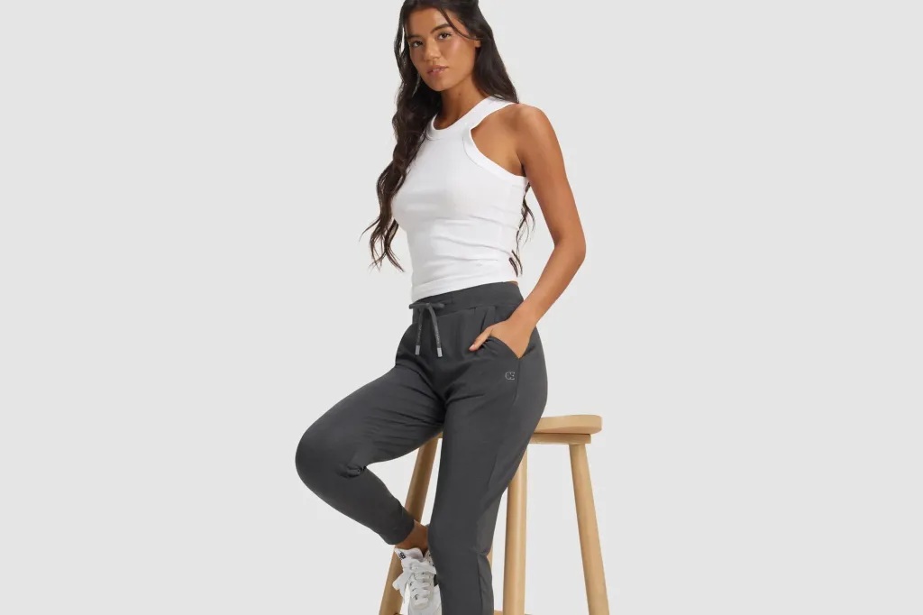 A woman with long dark hair wearing a white tank top and dark gray joggers with a logo.