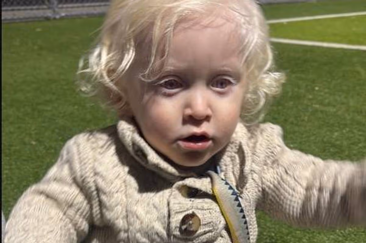 A young child with fair hair is standing on a grassy field, appearing to be in an outdoor setting. The child is dressed in a light-colored sweater and is looking towards the camera with an expression of curiosity. The backdrop features a partially visible sports field with markings, suggesting the child may be at a recreational facility or a sports event.