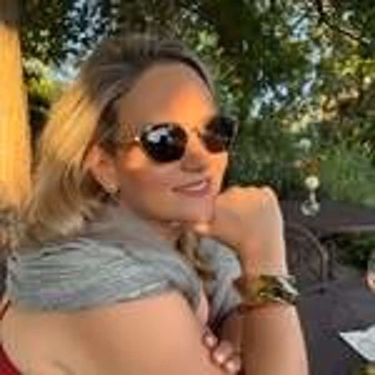An individual with long, light-colored hair is smiling and wearing sunglasses. They are seated outdoors, with a tree and foliage visible in the background.