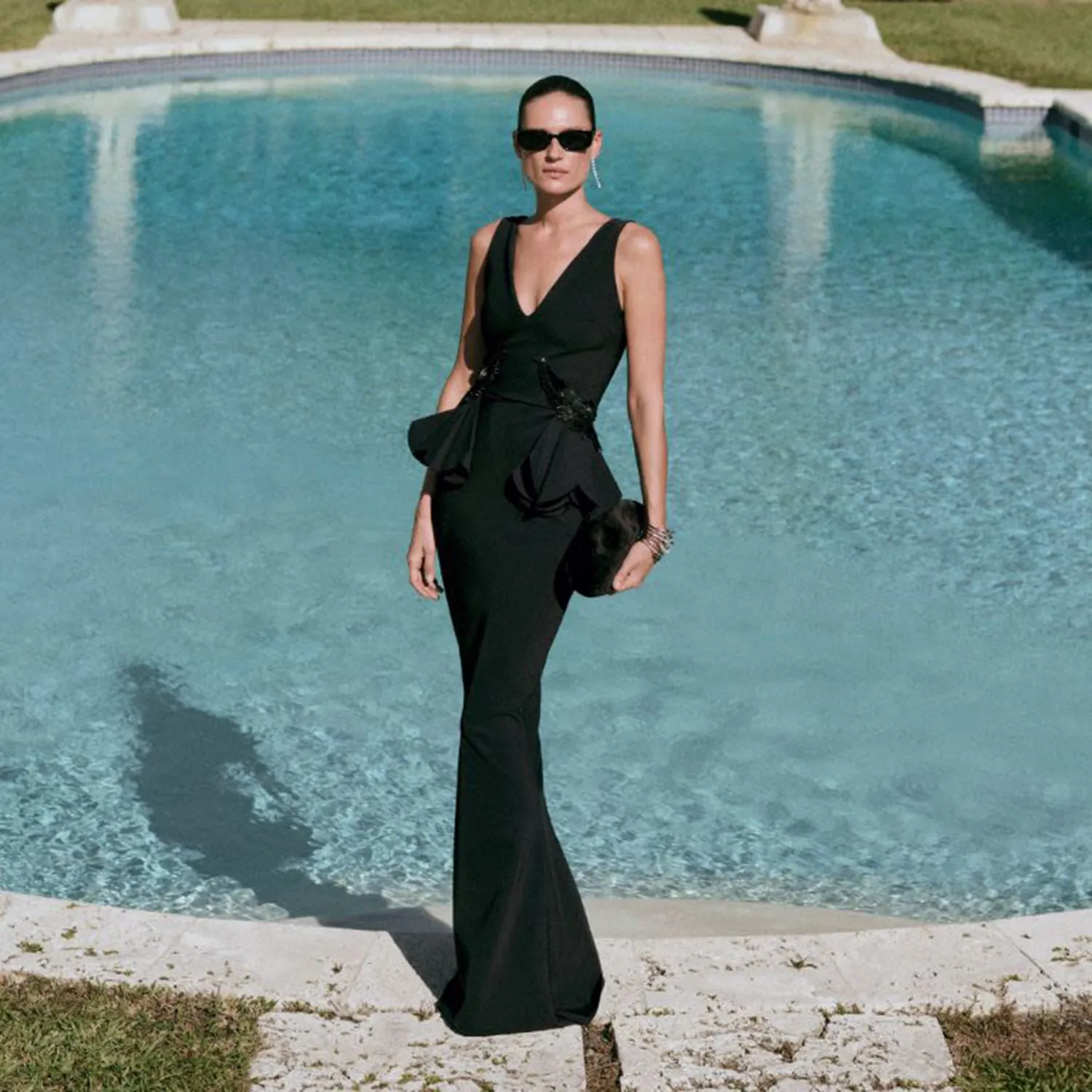Woman in a black peplum gown and sunglasses standing by a pool.