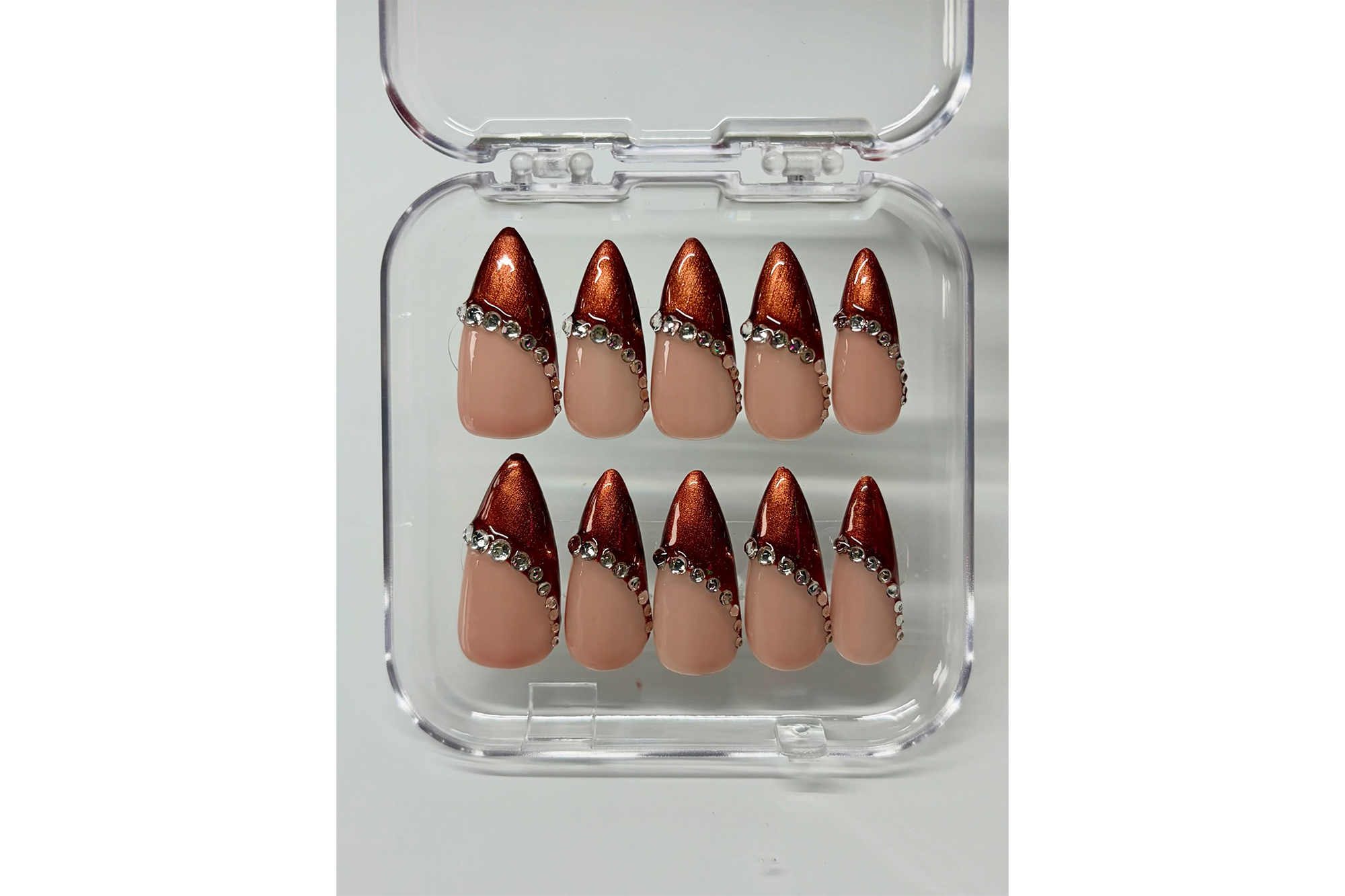 A set of almond-shaped press-on nails with nude bases, shimmering bronze tips, and rhinestone accents in a clear plastic case.