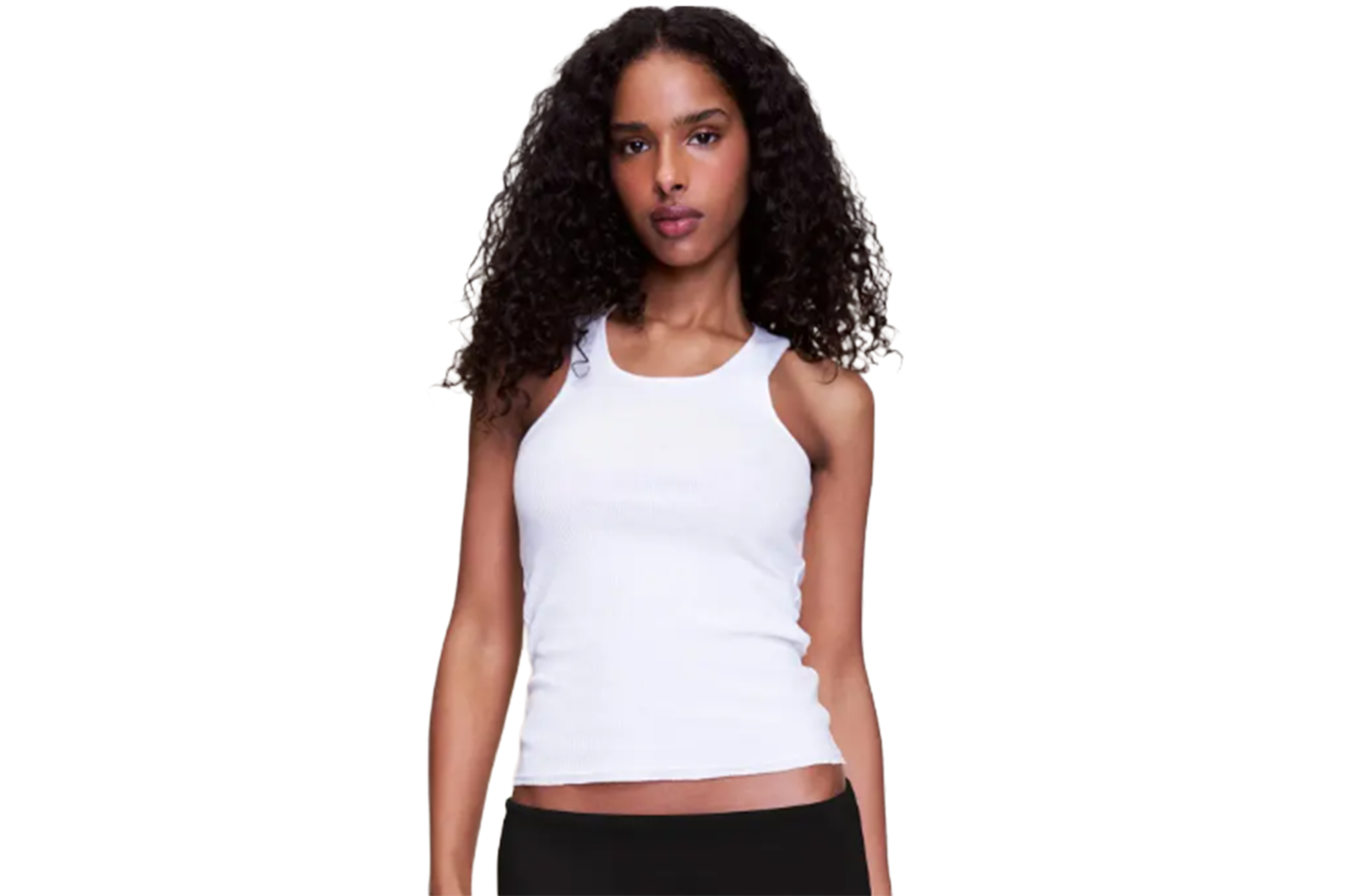 Woman with long, dark curly hair wearing a ribbed white tank top and black pants.