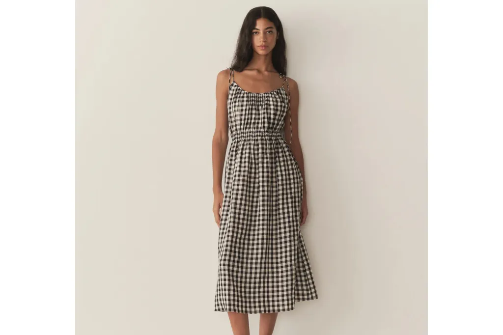 A woman with long dark hair stands facing the camera, wearing a black and white gingham midi dress with spaghetti straps and a shirred waist.