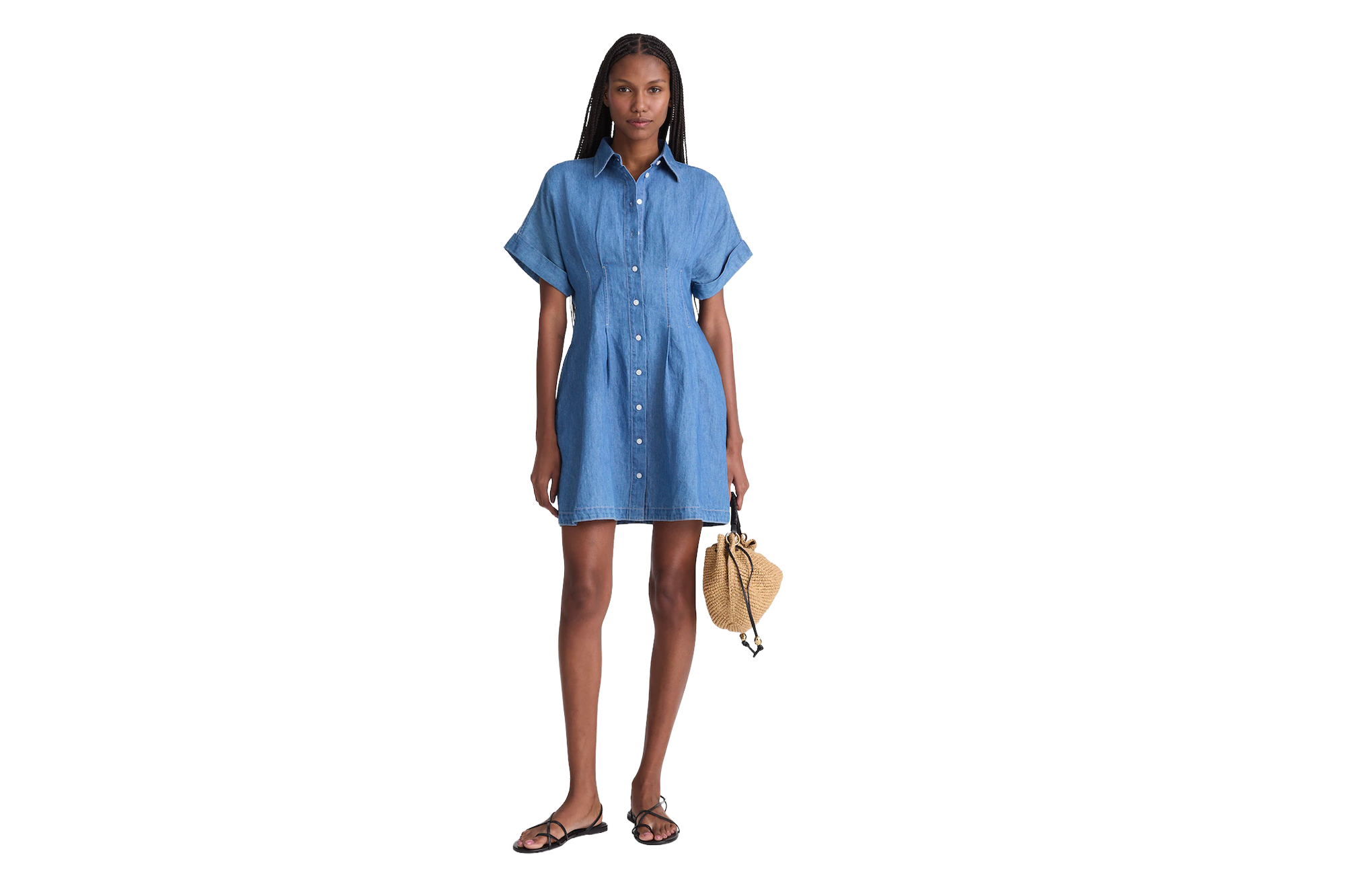 A model wearing a light-wash denim button-up mini dress, black flat sandals, and carrying a small straw bucket bag.