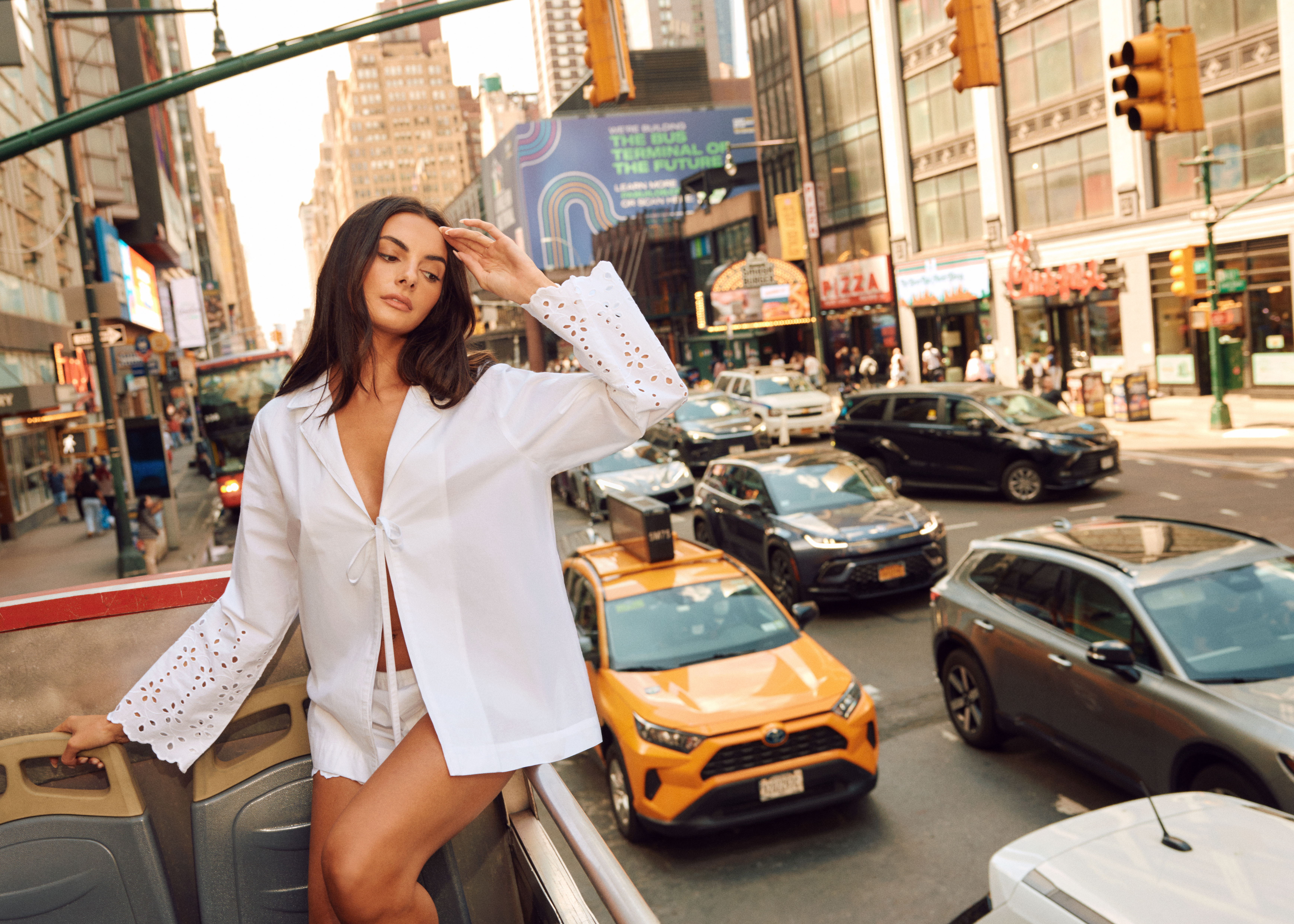 Paige De Sorbo in a white top standing on an open-top bus on a busy street in New York City.