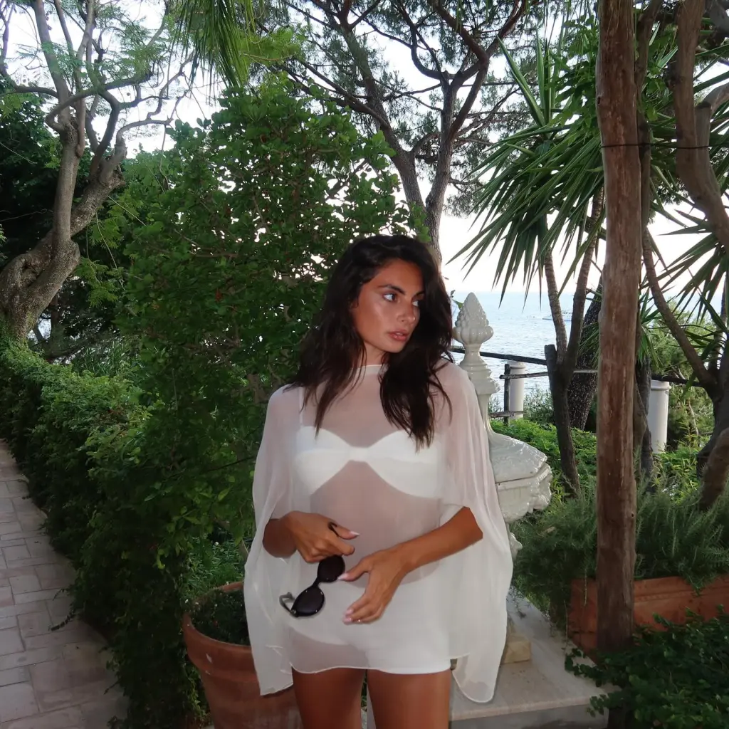 Paige DeSorbo wearing a sheer white kaftan over a white bikini, holding sunglasses.
