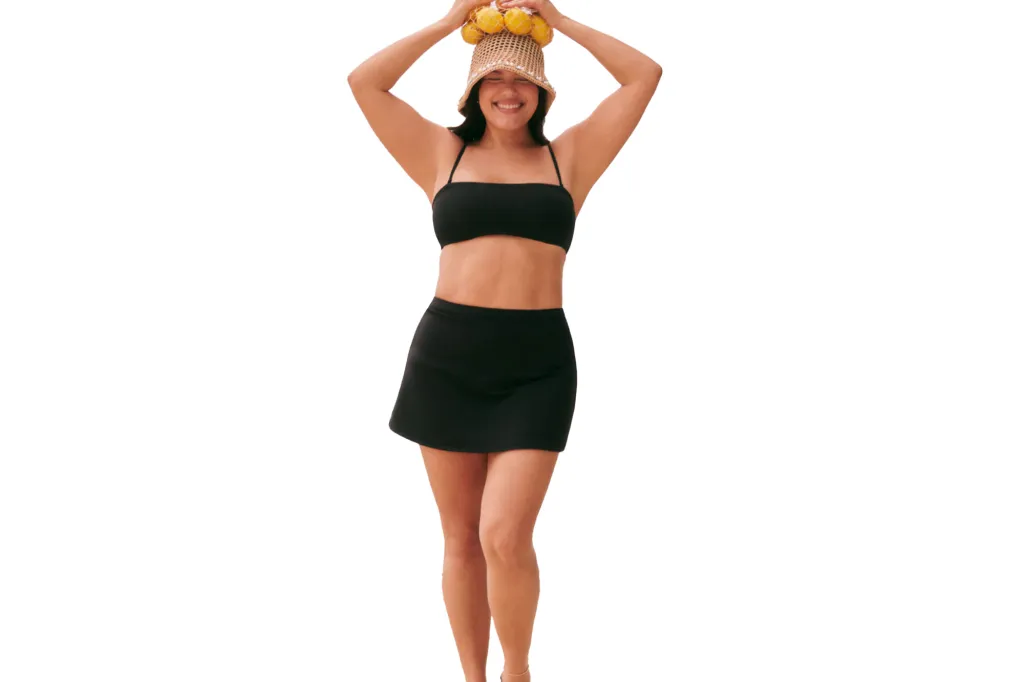 A woman in a black swimsuit and black swim skirt balancing a woven straw hat filled with lemons on her head.