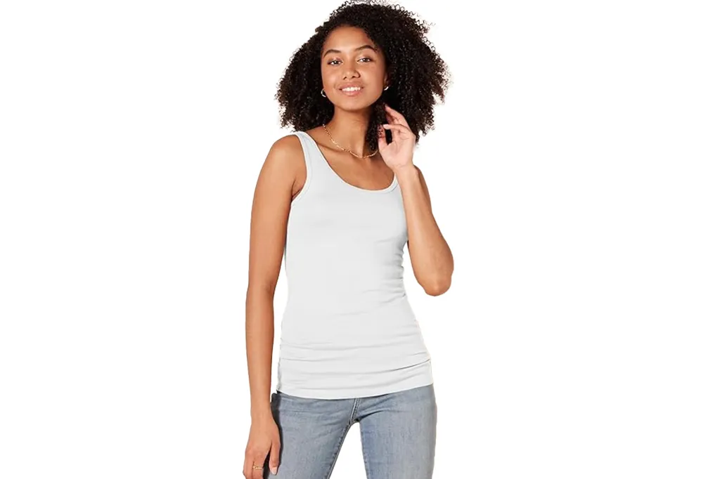 A woman wearing a white tank top and blue jeans, with one hand near her face.