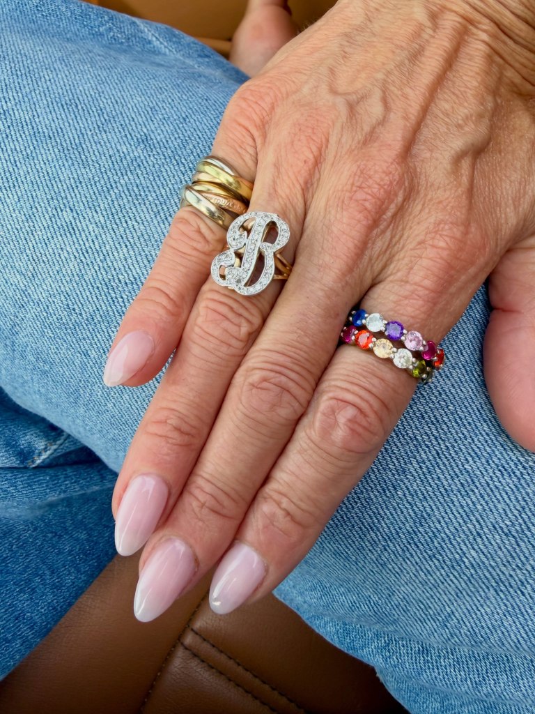 Hand wearing a ring with a diamond 'B' on the middle finger and two multi-gemstone rings on the ring finger.