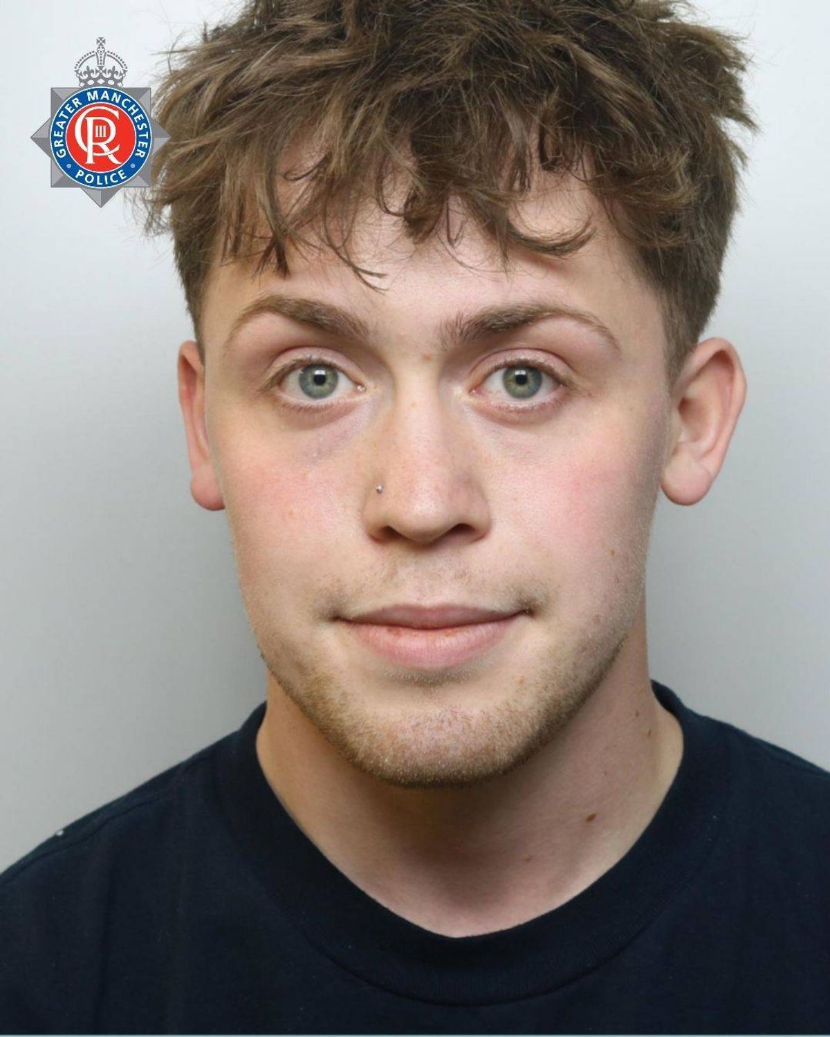 A formal portrait of a young individual with short hair, wearing a black top, and displaying a neutral expression against a plain background. The individual has light-colored eyes and a clean-shaven face.