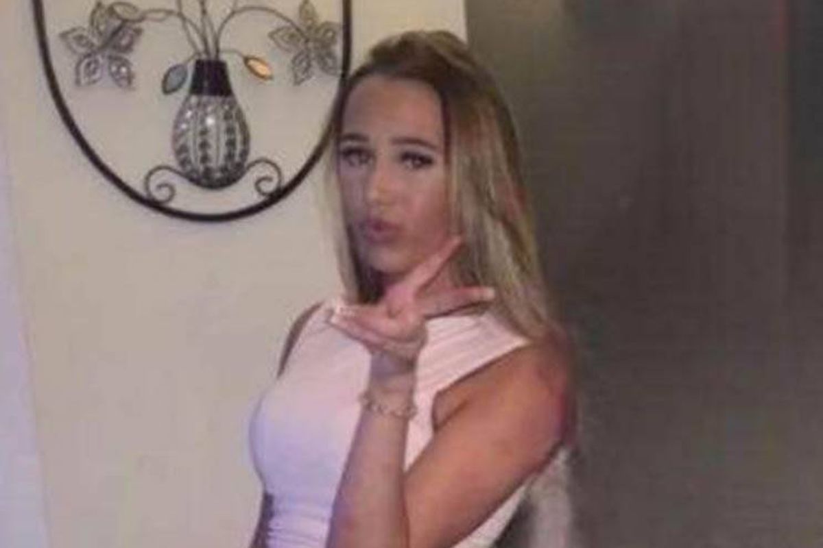 An individual with blonde hair is standing in front of a white wall adorned with a decorative circular art piece. The person is dressed in a sleeveless white garment and is making a peace sign with their hand.